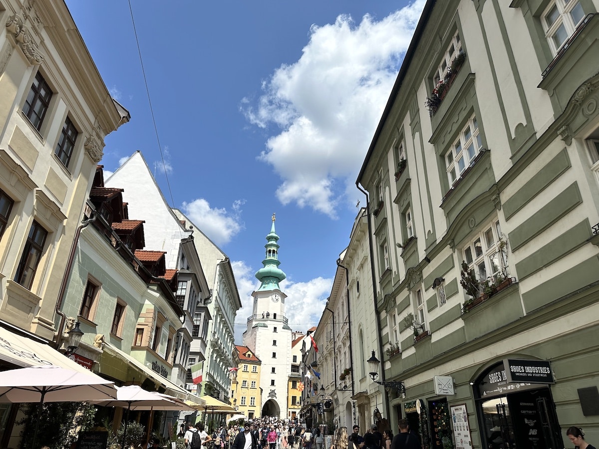 Blick auf eine Straße mit historischen Gebäuden und einem markanten Turm unter einem blauen Himmel mit Wolken.