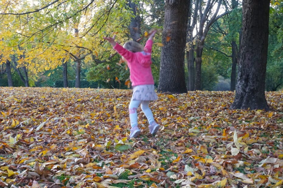 Ein Mädchen in einem pinken Pullover springt in einem Laubhaufen im Herbst.