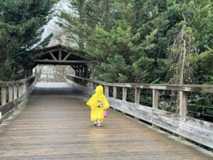 Ein Kind in einem gelben Regenmantel geht über eine Holzbrücke in einem bewaldeten Bereich.