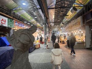 Ein Markt mit Verkaufsständen und einer Statue im Vordergrund, die eine Person darstellt. Menschen gehen durch den Markt.