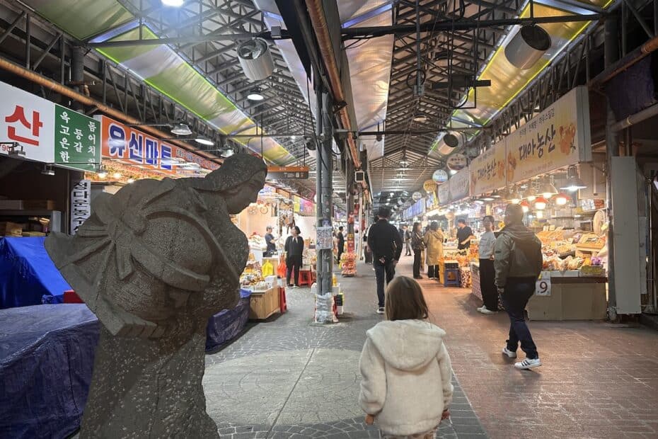 Ein Markt mit Verkaufsständen und einer Statue im Vordergrund, die eine Person darstellt. Menschen gehen durch den Markt.