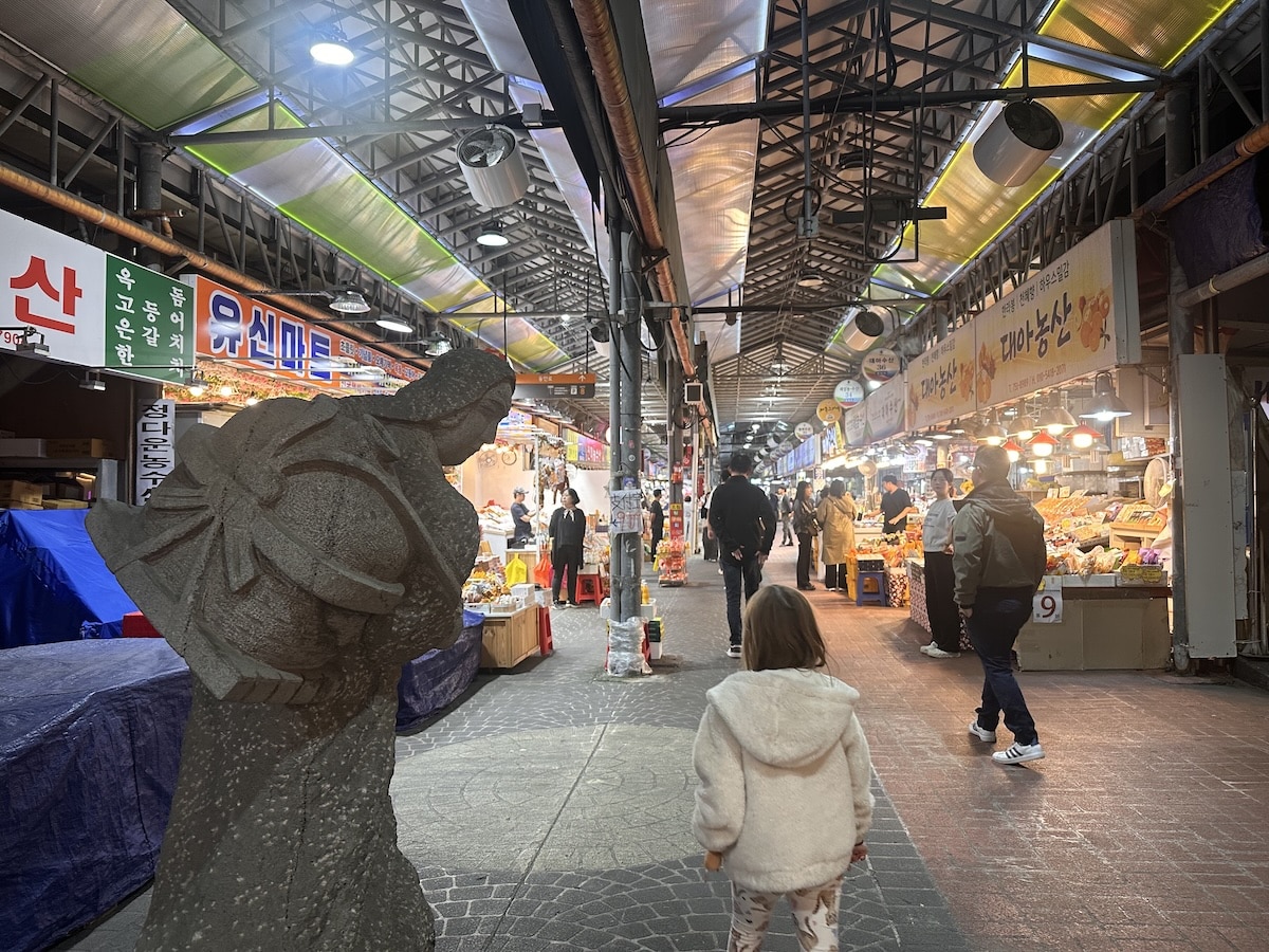 Ein Markt mit Verkaufsständen und einer Statue im Vordergrund, die eine Person darstellt. Menschen gehen durch den Markt.