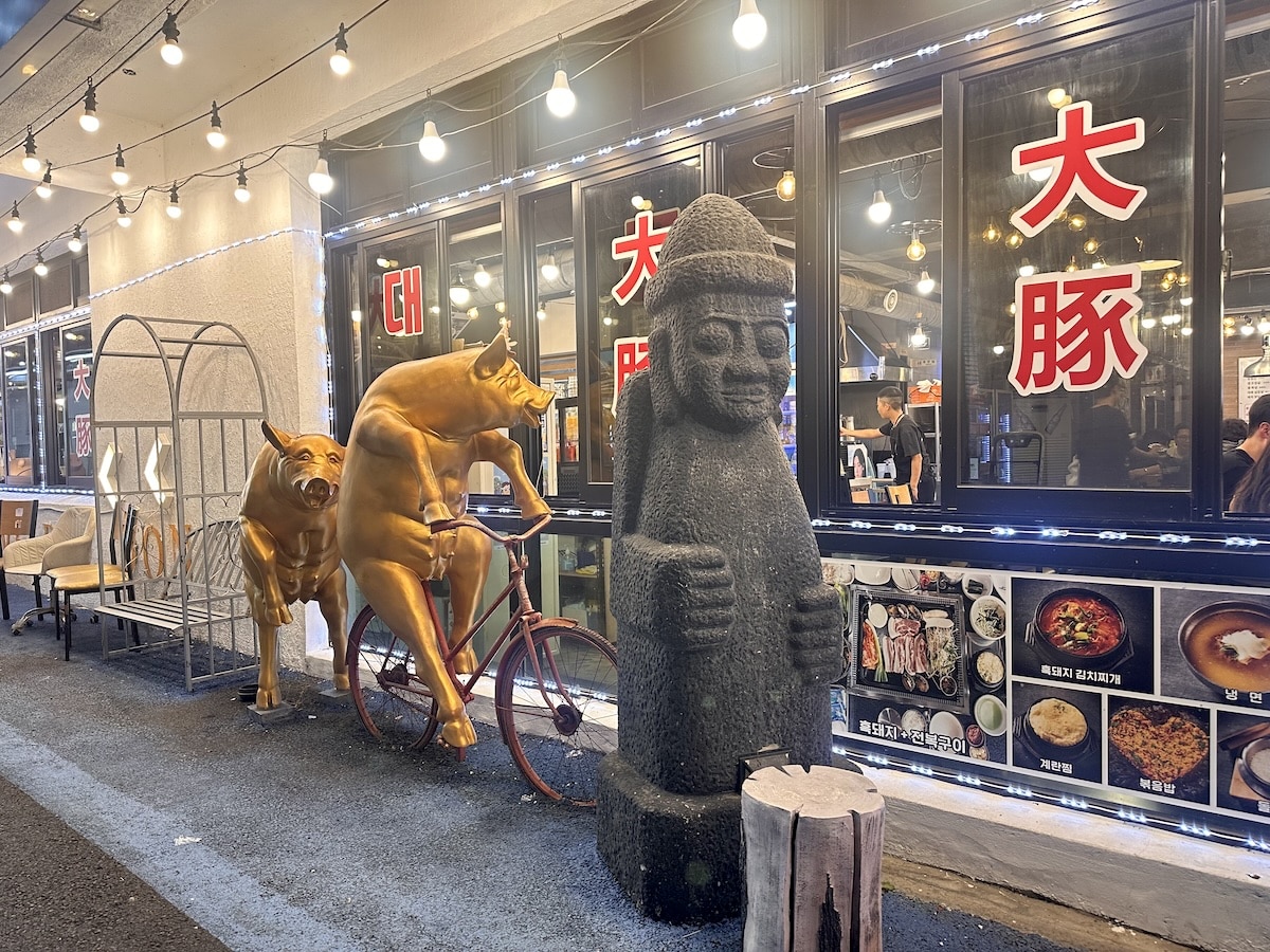 Goldene Stier-Skulpturen und eine Steinfigur vor einem Restaurant mit beleuchteten Schildern.JPork Street Jeju
