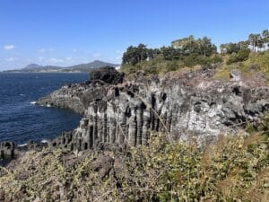 Felsenformation mit Säulenstruktur an der Küste, umgeben von Wasser und Vegetation.