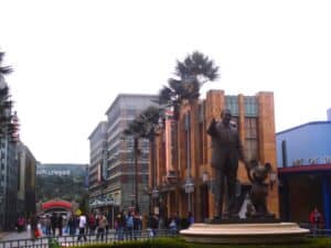 Bronzestatue von Walt Disney, der mit Mickey Mouse in einem Freizeitpark steht, umgeben von Gebäuden und Palmen.