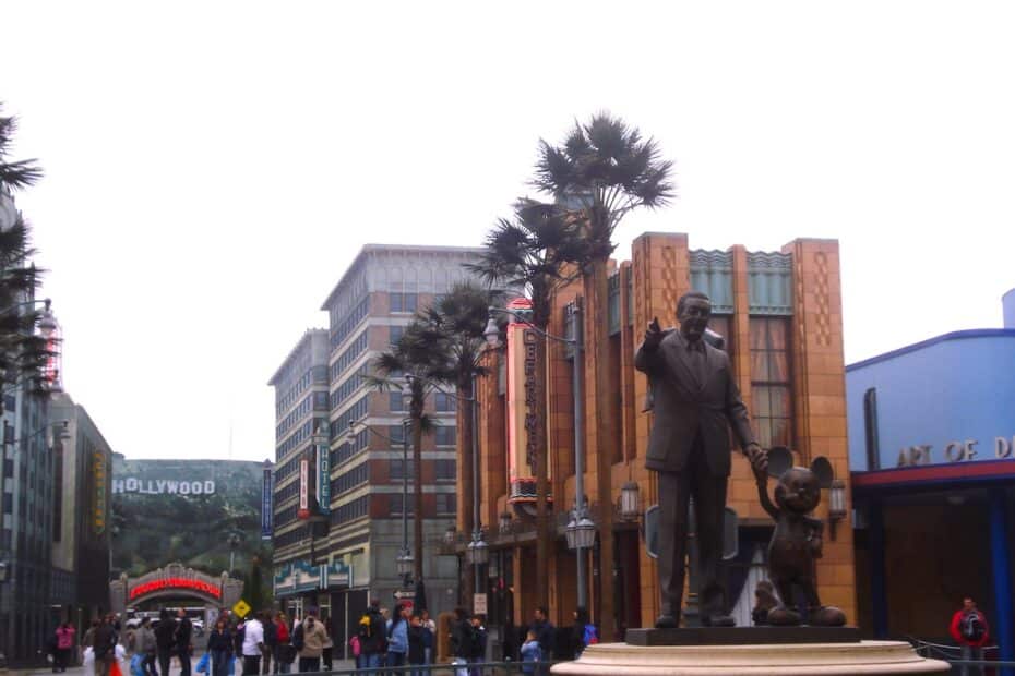 Bronzestatue von Walt Disney, der mit Mickey Mouse in einem Freizeitpark steht, umgeben von Gebäuden und Palmen.