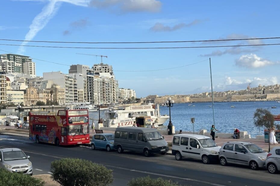 Roter Doppeldeckerbus fährt entlang der Küste mit Blick auf das Wasser und die Stadtarchitektur im Hintergrund.