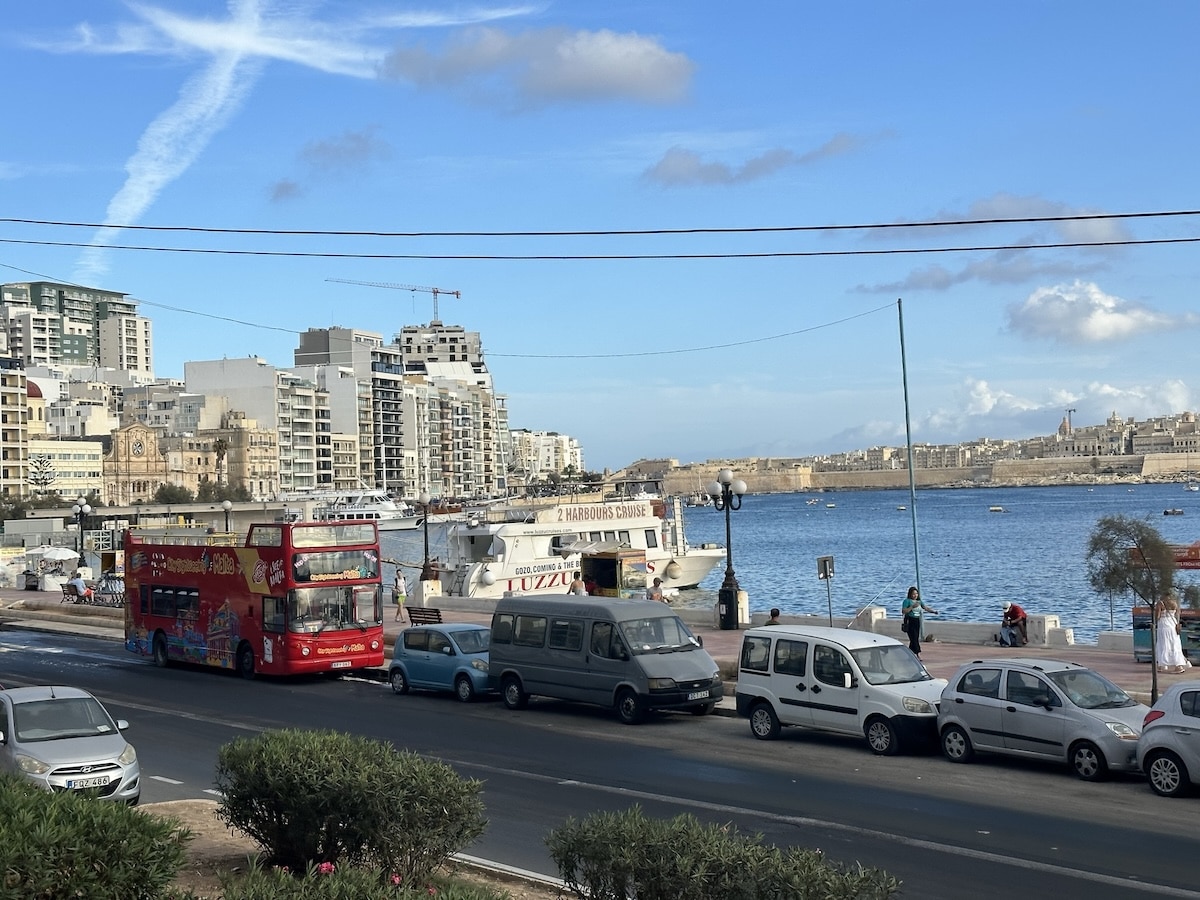 Roter Doppeldeckerbus fährt entlang der Küste mit Blick auf das Wasser und die Stadtarchitektur im Hintergrund.