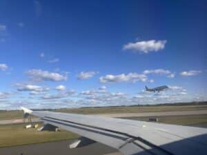 Flugzeugflügel mit Blick auf ein landendes Flugzeug vor einem blauen Himmel mit Wolken.