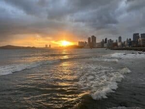 Sonnenuntergang, der über einer Küstenstadt mit Wolken und Wellen am Wasser sichtbar ist.