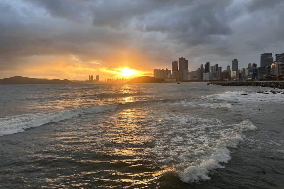 Sonnenuntergang, der über einer Küstenstadt mit Wolken und Wellen am Wasser sichtbar ist.