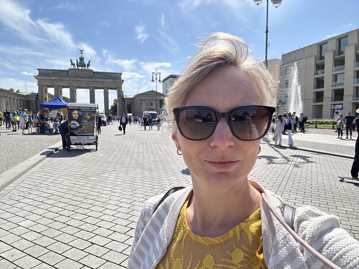 Frau mit Sonnenbrille und gelbem Oberteil steht vor dem Brandenburger Tor in Berlin an einem sonnigen Tag.