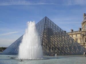 Glaspyramide des Louvre mit einem Wasserbrunnen im Vordergrund, umgeben von klarem Himmel und architektonischen Details des Museums.