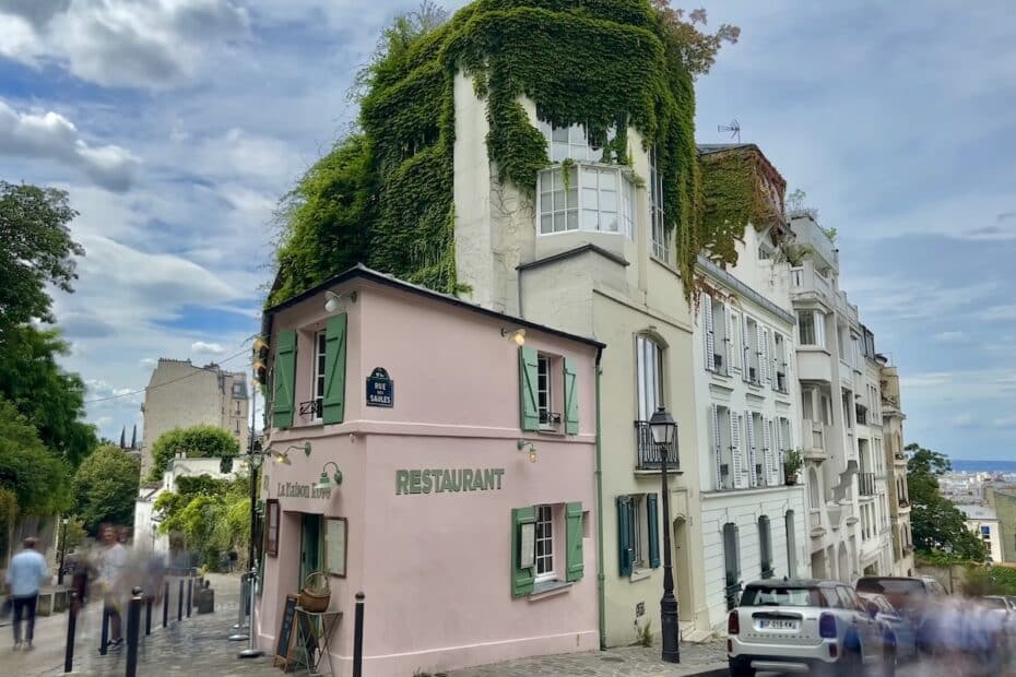Ein pinkes Restaurant mit grüner Bepflanzung auf dem Dach, umgeben von weißen Gebäuden in einer urbanen Umgebung.