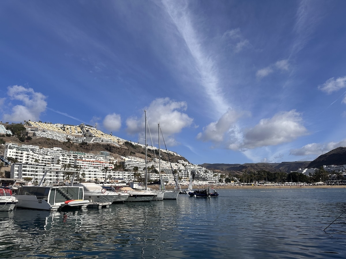 Blick auf den Hafen von Puerto Rico auf Gran Canaria mit Booten, Uferhäusern am Hang und blauem Himmel – Landschaftsaufnahme aus einer Küstenstadt.