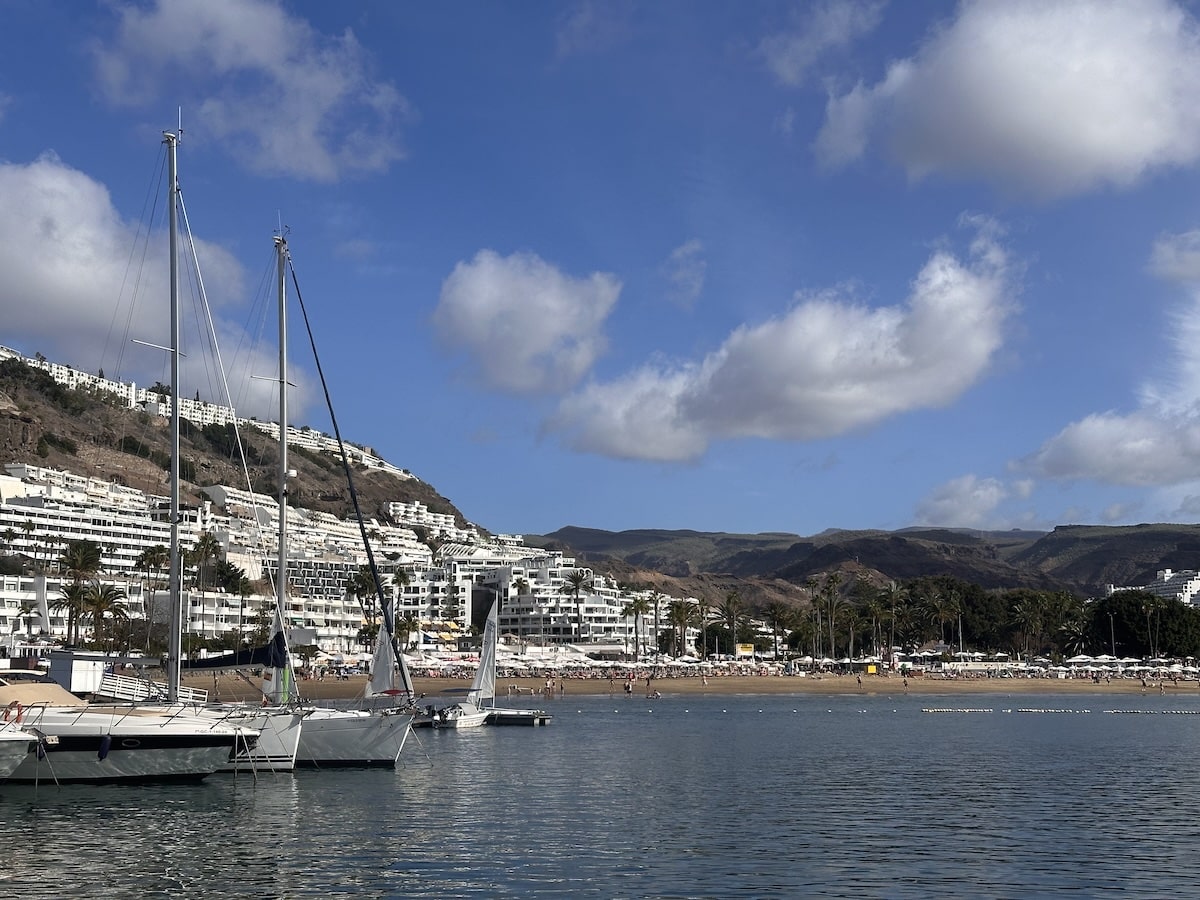 Blick aufs Hafenviertel von Gran Canaria mit Segelbooten im Wasser, weiße Apartmentgebäude an einer Küstenlinie und einem blauen Himmel; Bezug zu Puerto de Mogán und Gran Canaria wird erkennbar.