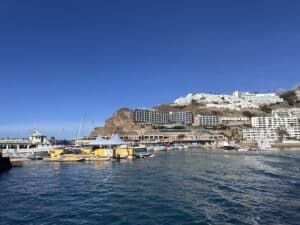 Hafenansicht von Gran Canaria in Puerto Rico mit Uferpromenade, vielen Booten, weißen Hotelbauten am Hang, klarem blauen Himmel und ruhigem Wasser