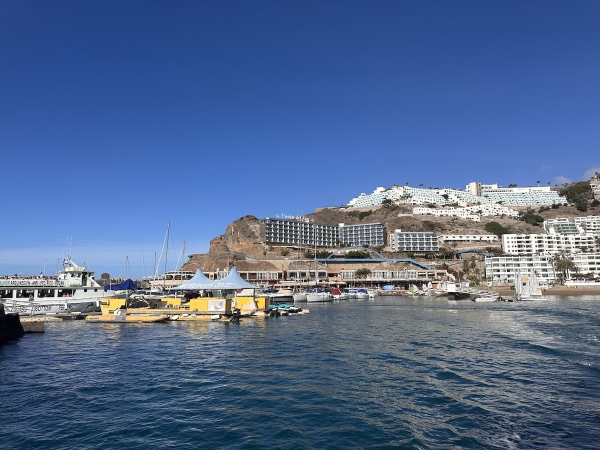 Hafenansicht von Gran Canaria in Puerto Rico mit Uferpromenade, vielen Booten, weißen Hotelbauten am Hang, klarem blauen Himmel und ruhigem Wasser