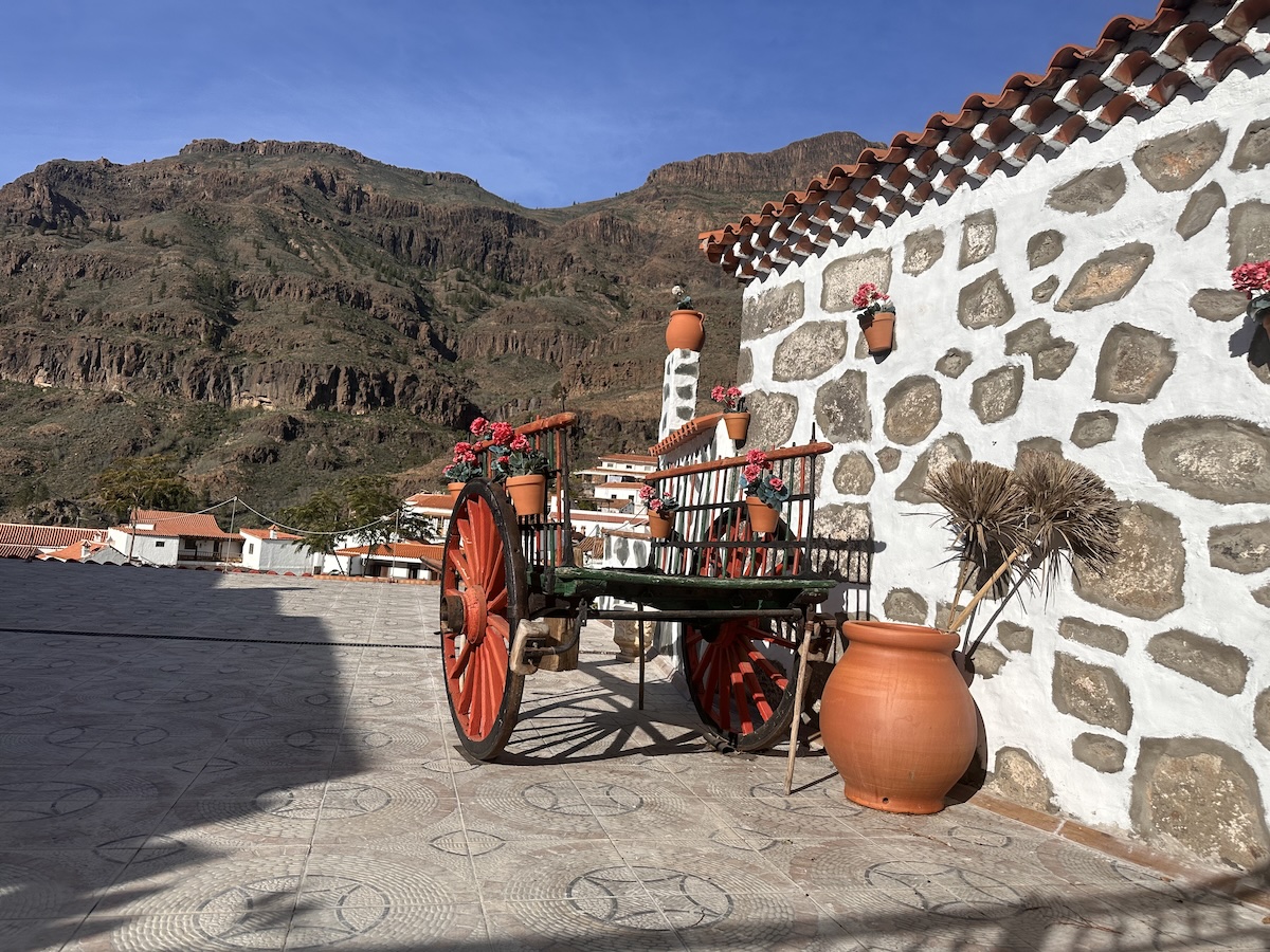 Gran Canaria Inselinneres: traditioneller roter Holzwagen vor einer Steinmauer mit Terracotta-Töpfen, imposante Berglandschaft, klare blaue Himmelsfläche und malerische Dörfer im Hintergrund.
