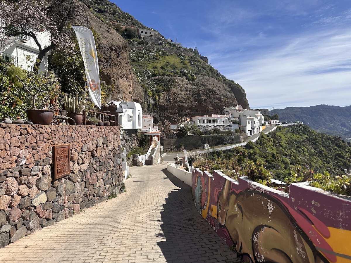 Malerischer Weg, Inselinneres mit weißen Häusern, Steinmauern und einer graffitiartigen Mauer im Vordergrund; bergige Küstenlandschaft erstreckt sich im Hintergrund.