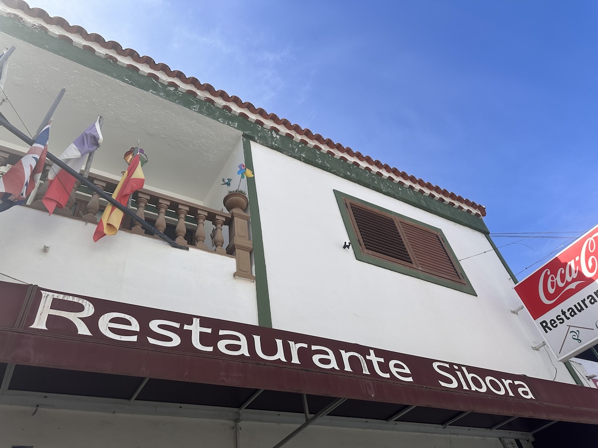 Restaurante Sibora mit bunten Flaggen am Balkon, klarer blauer Himmel und eine einladende Fassade – typisches kanarisches Restaurant-Ambienteum mittag