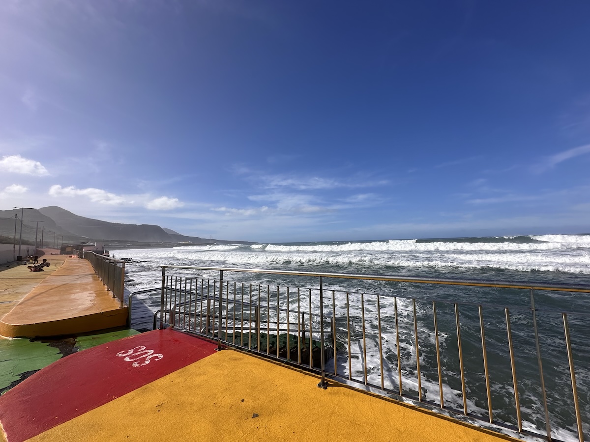 Klares Blau des Himmels über dem Meer, roter-ockerfarbener Boden am Vordergrund mit Gelb- oder Orangetönen, ruhige Küstenpromenade und brechende Wellen – , Roque Bentayga und Tejeda bleiben im Hintergrund sichtbar.