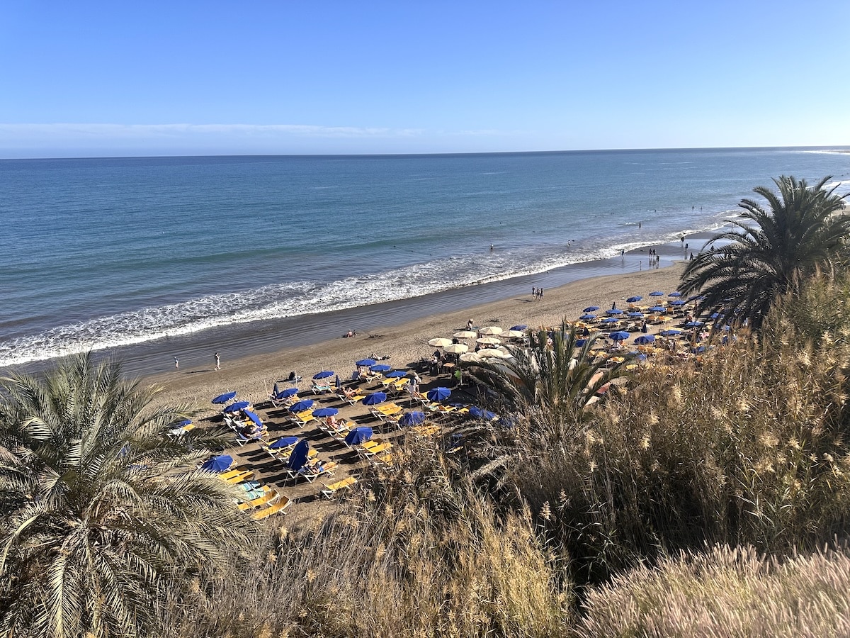 Strandbild auf Gran Canaria mit goldenem Sand, blauen Sonnenschirmen und Palmen am Vordergrund, Blick aufs Meer; Ort nahe Maspalomas oder Playa del Inglés im Süden der Insel Gran Canaria.