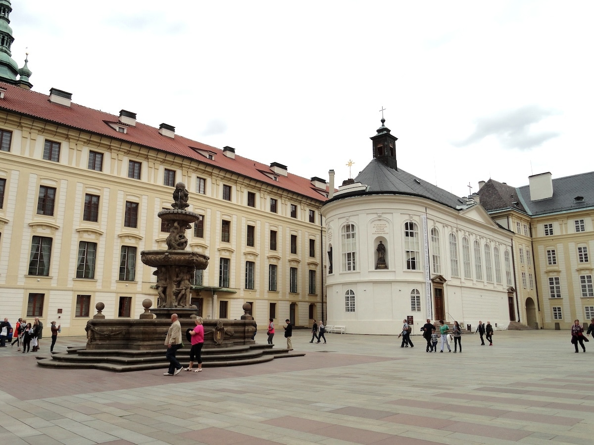 Weiträumiger Innenhof der Prager Burg mit Springbrunnen, klassizistischen Fassaden und rotbraunem Ziegeldach in Prag bei grauem, bewölktem Himmel.