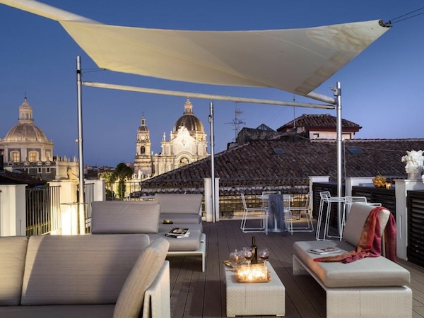 Ruhige Rooftop-Terrasse mit Sitzgelegenheiten und warmen Lichtern, dahinter der Blick auf den Piazza del Duomo in Catania bei blauem Abendhimmel.