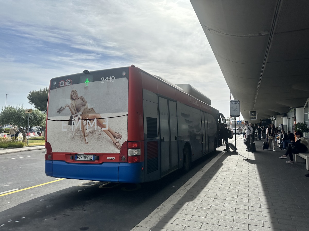 Blauer und roter Bus an einem Bahnsteig, Menschen an der Station und ein Werbeaufkleber am Heck vermitteln Ankommen in Catania