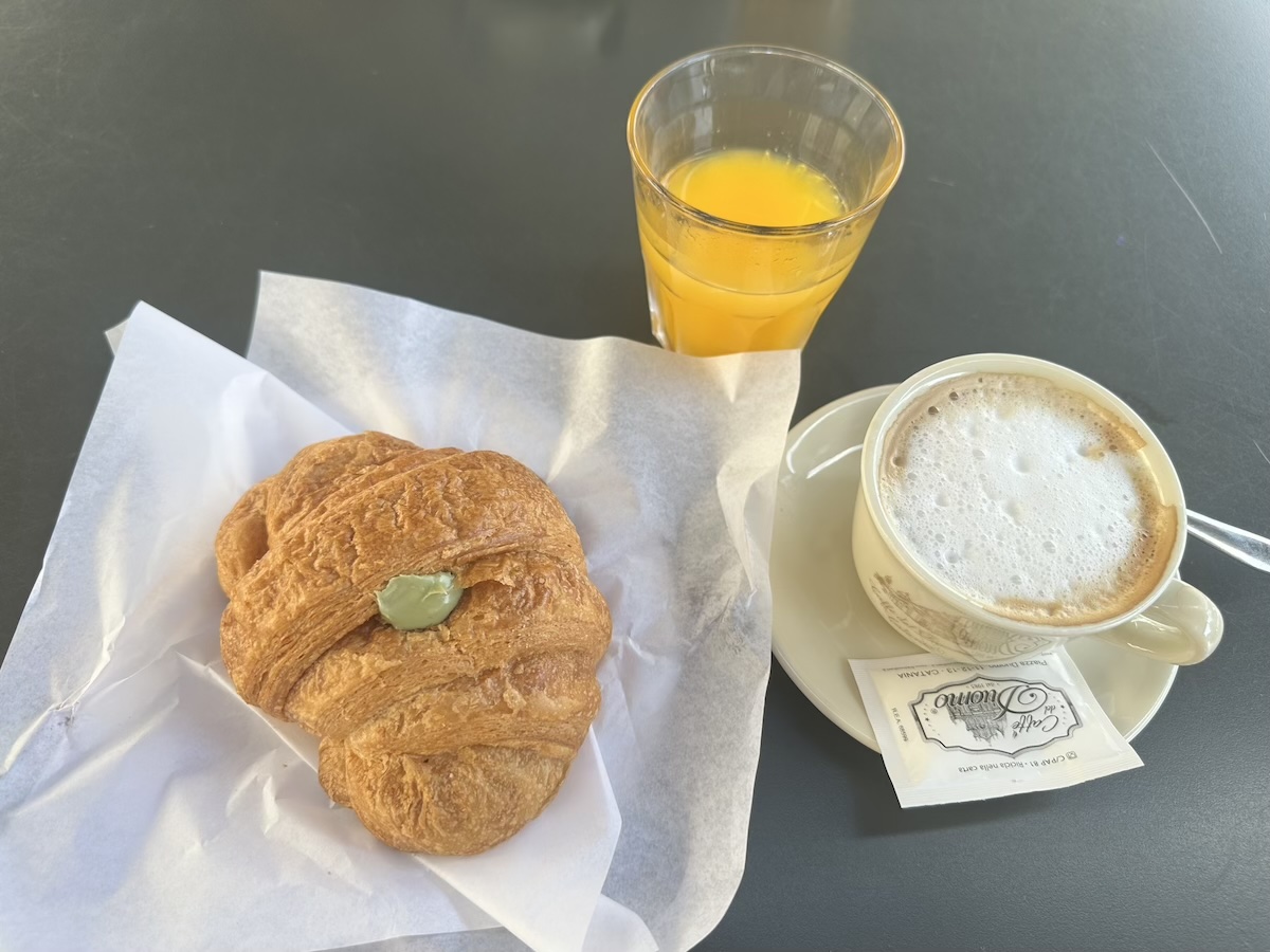 Goldbraunes Brioche liegt auf Papier, dazu ein orangefarbener Drink und ein schaumiger Kaffee – typisch sizilianisches Streetfood aus Catania.