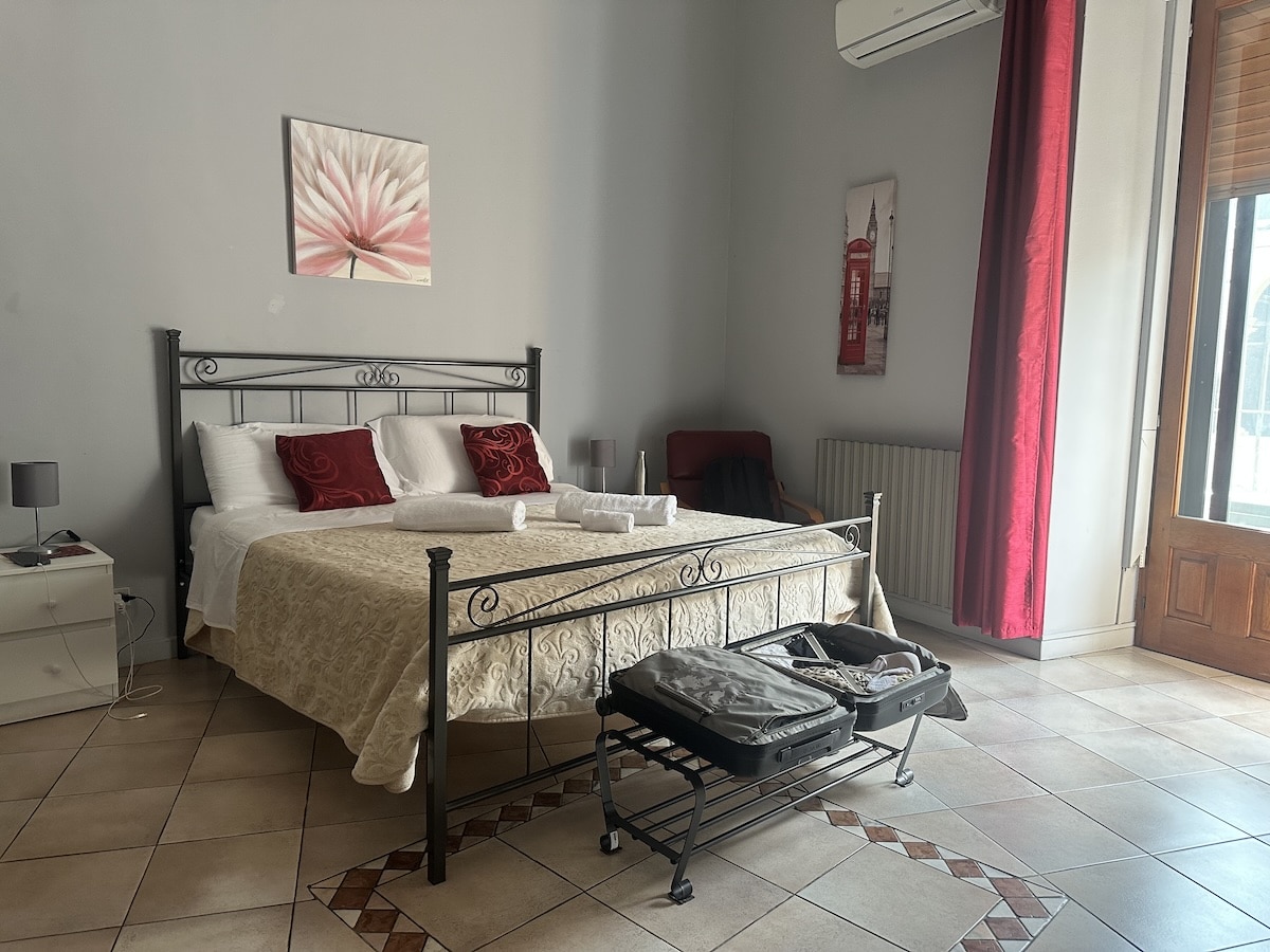 Ruhiges Schlafzimmer mit weißem Bettgestell, rotem Vorhang am Fenster und gefliesten Böden, passend zu Catania Sehenswürdigkeiten & Tipps für deinen Besuch.