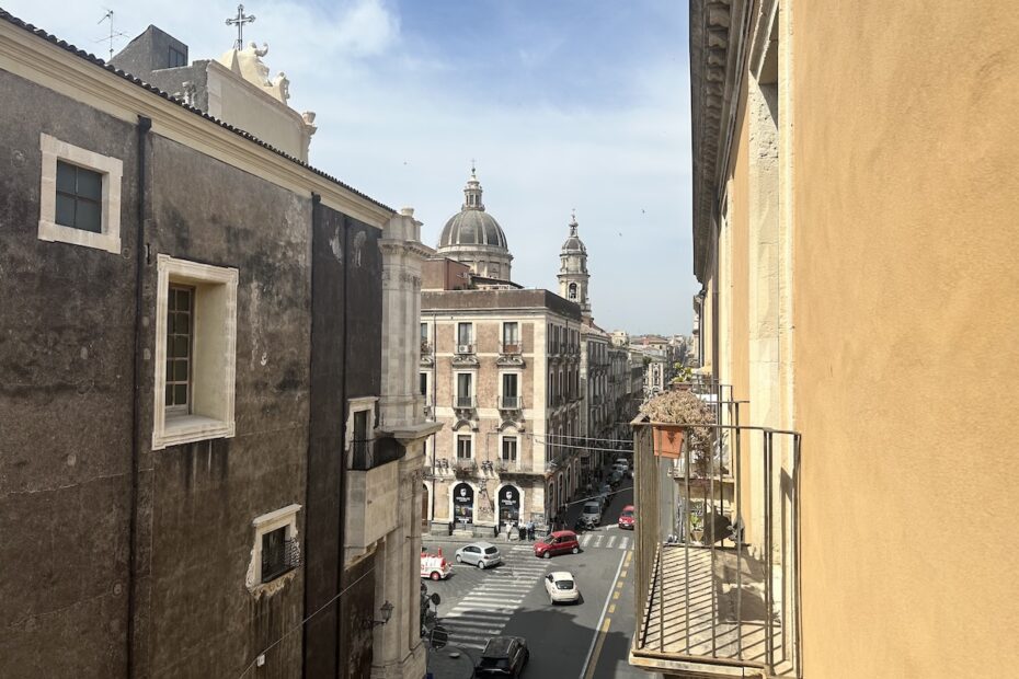 Blick aus der Unterkunft Terrazza Santa Chiara in Catania: die Kuppel von Piazza del Duomo über der Straße an der Via Etnea, mit beige-grauen Fassaden und Autos.