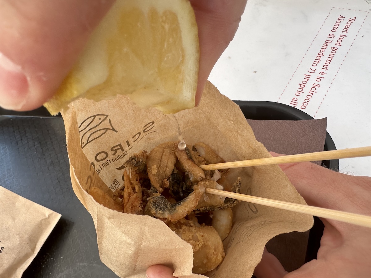 Hand mit Stäbchen hält ein Cartoccio di Pesce: goldbraune, knusprig-frittierte Fischstücke im Papiertütchen vor dem Fischmarkt La Pescheria in Catania.