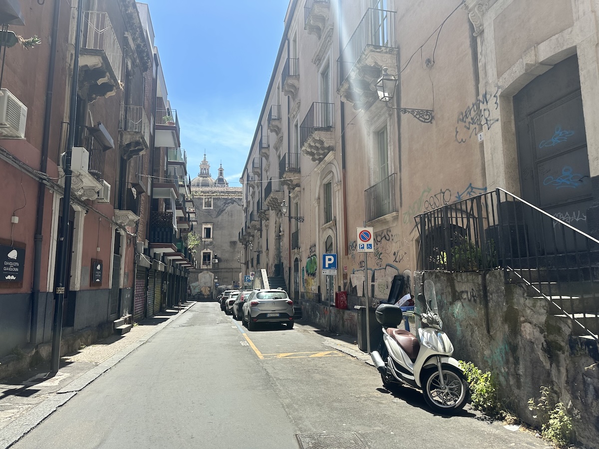 In Catania führt eine enge Gasse durch die Altstadt in Sizilien zwischen mehrstöckigen Fassaden, graffitibeschmierten Wänden und geparkten Rollern, mit sichtbaren Schildern.