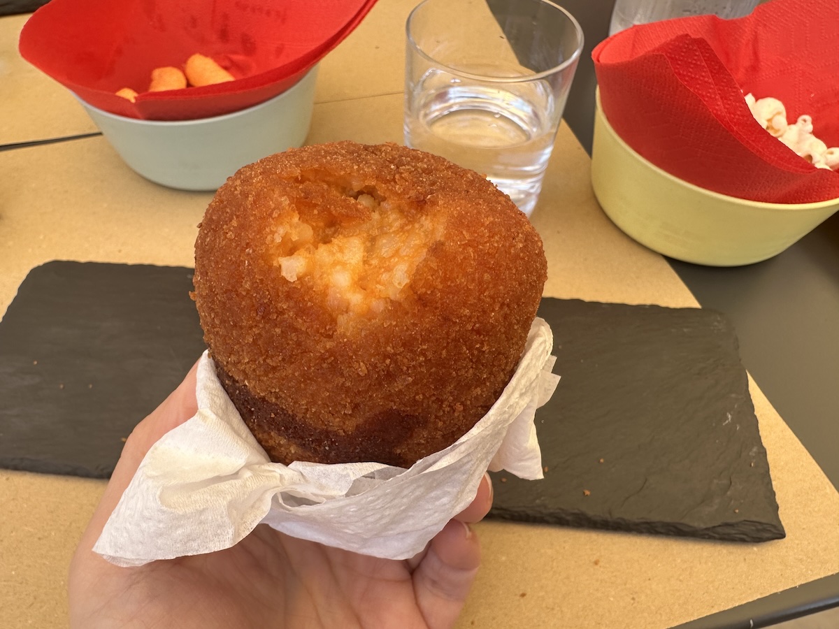 Goldbraunes, rund frittiertes Arancini in einer Hand mit Krümeln auf Backpapier, im Hintergrund rote Schalen und ein Glas Wasser