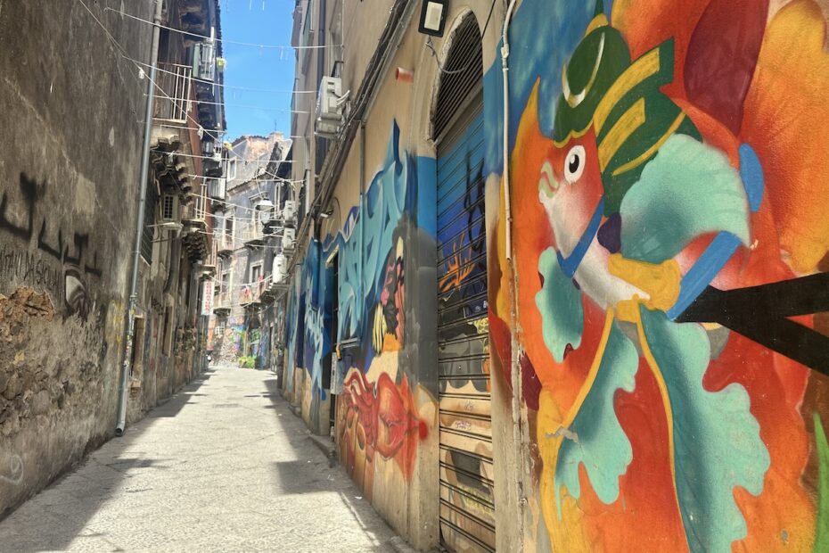 In Catania im Viertel San Berillo führt eine enge Gasse zwischen Hauswänden mit farbigem Graffiti und Rolläden in Richtung Tageslicht, darunter wirkt der Weg eher rau.