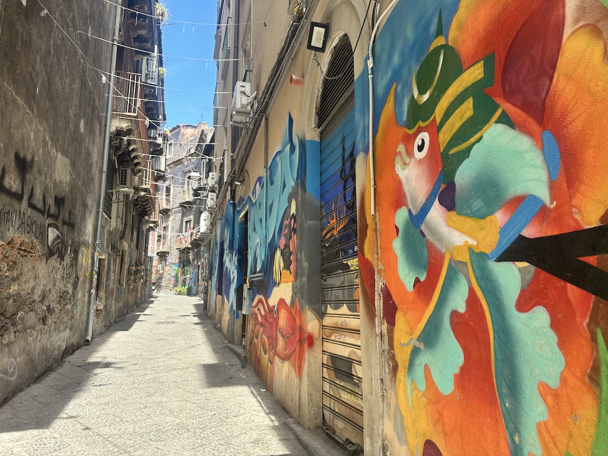 In Catania im Viertel San Berillo führt eine enge Gasse zwischen Hauswänden mit farbigem Graffiti und Rolläden in Richtung Tageslicht, darunter wirkt der Weg eher rau.