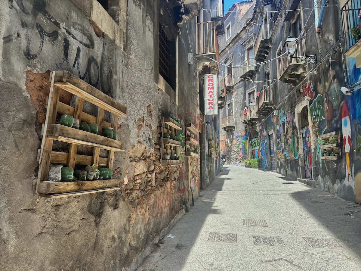 Enge graue Gasse in Catania mit buntem Streetart an den Wänden, Läden mit Kisten und Gläsern sowie einem roten Schild mitten im Viertel
