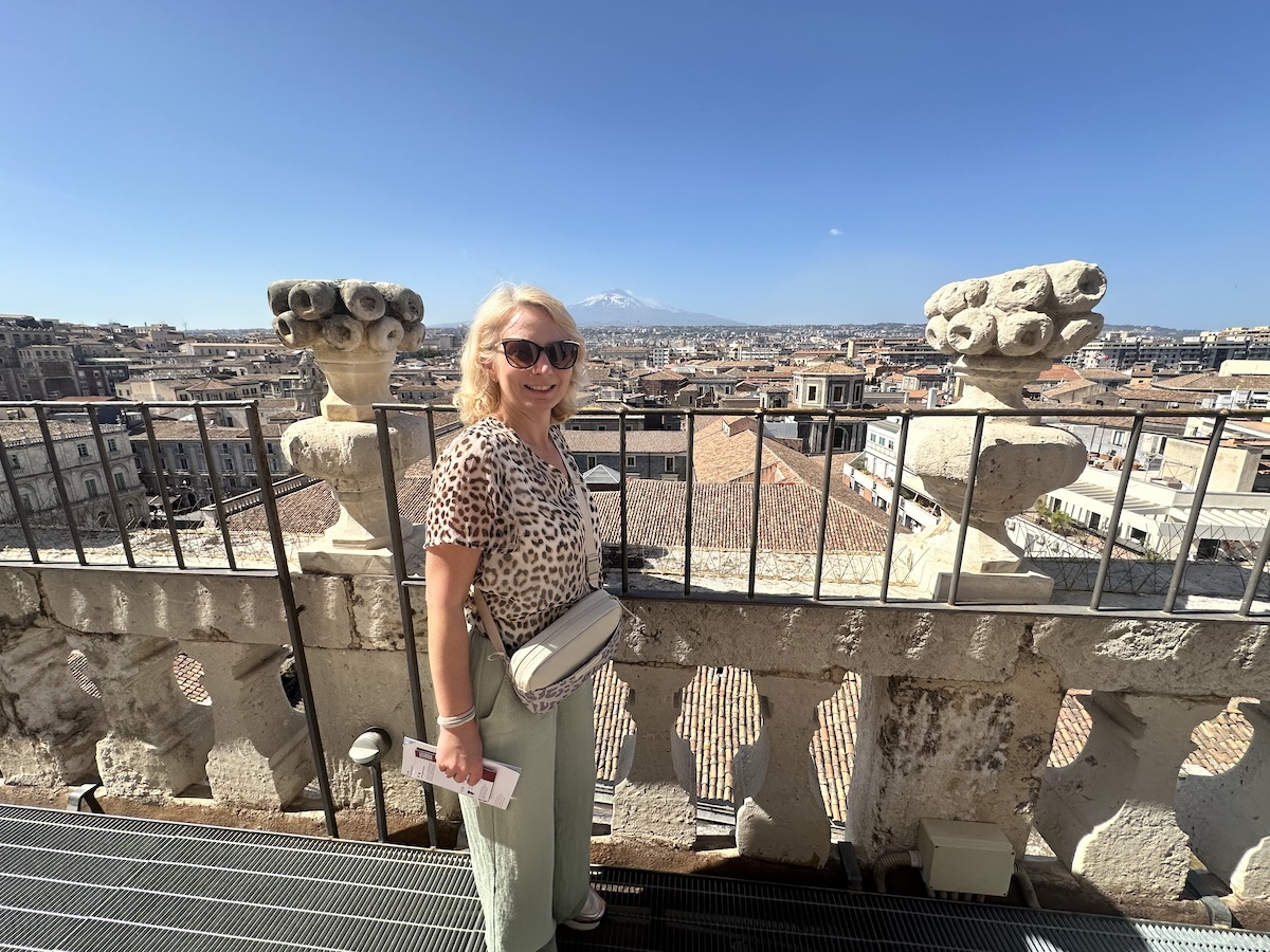 Auf der steinernen Terrasse in Catania steht eine Reisende im leopardfarbenen Outfit, blickt über Dächer mit dem Ätna im Hintergrund in den blauen Himmel.