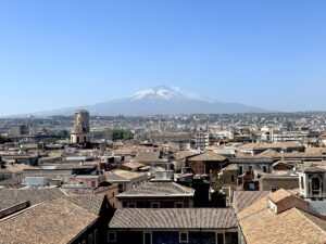 Blick über Catania mit der Kathedrale von Sant’Agata und roten Dächern, im Hintergrund der Ätna mit schneebedecktem Gipfel unter blauem Himmel.
