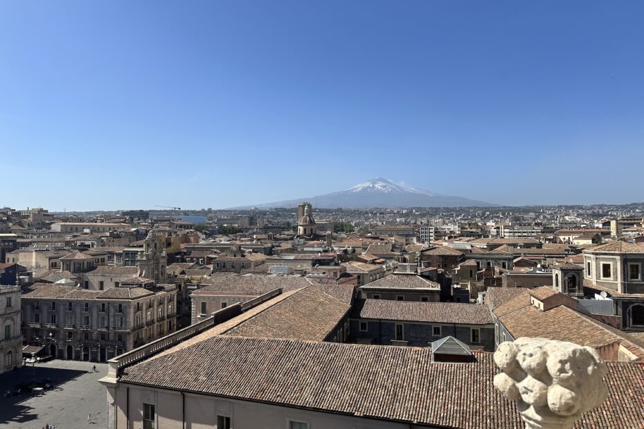 Blick über Catania mit roten Ziegeldächern und dem schneebedeckten Ätna am Horizont, klarer Himmel und typisch sizilianische Altstadt-Atmosphäre für deinen Tag.
