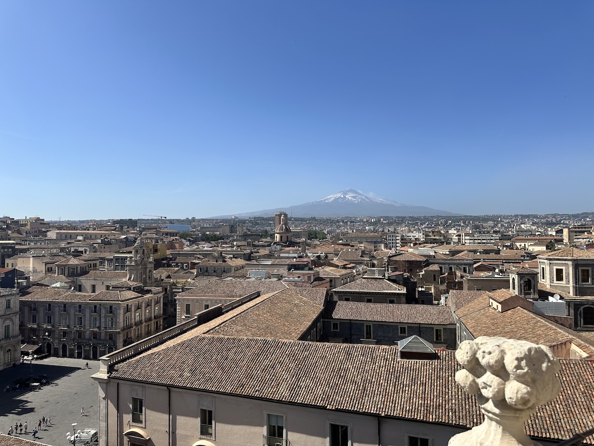 Blick über Catania mit roten Ziegeldächern und dem schneebedeckten Ätna am Horizont, klarer Himmel und typisch sizilianische Altstadt-Atmosphäre für deinen Tag.
