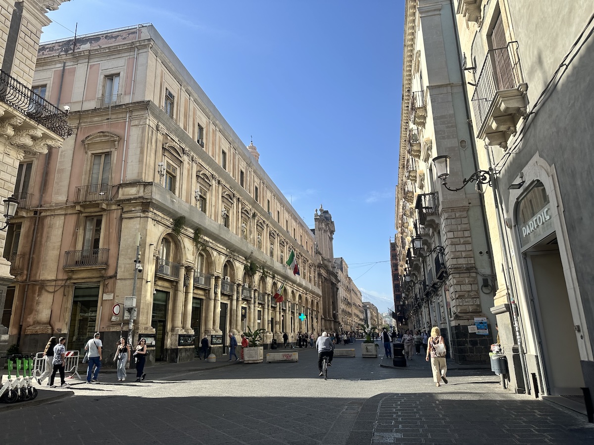 Catania Altstadt mit beigen Sandsteinfassaden und Arkaden an einer breiten Straße, Passanten und Fahnen im klaren Himmel – Catania Sehenswürdigkeiten.