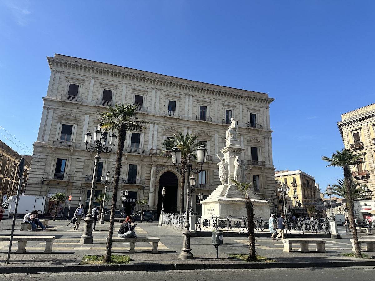 Weiße Treppen und Statuen vor dem Teatro Romano in Catania unter blauem Himmel mit Palmen und klassischer Fassade für deinen Tagesplan bei Catania an einem Tag.