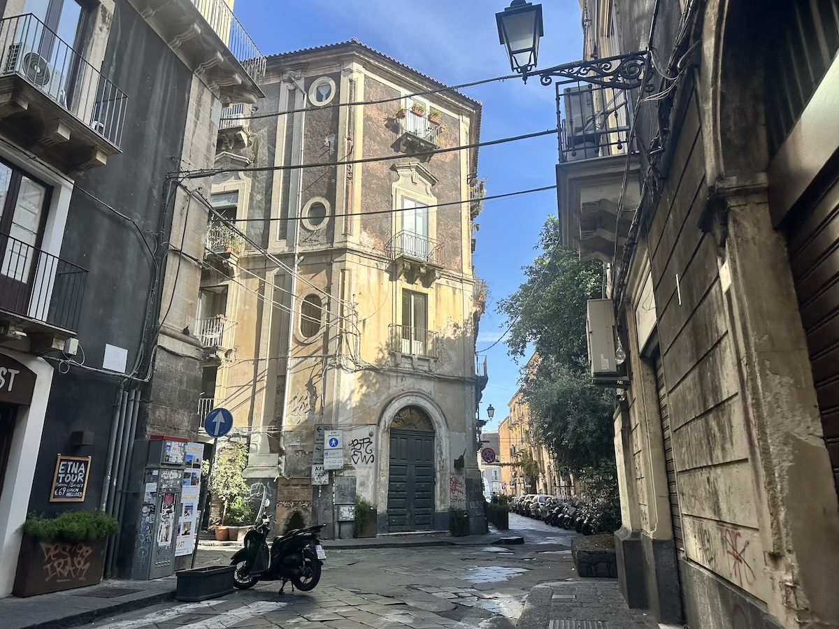 Enge Gasse in Catania mit barocken Fassaden und schattiger Stimmung, Straßenschild „Ätna Tour“ und Blick nach vorn zwischen Gebäuden.