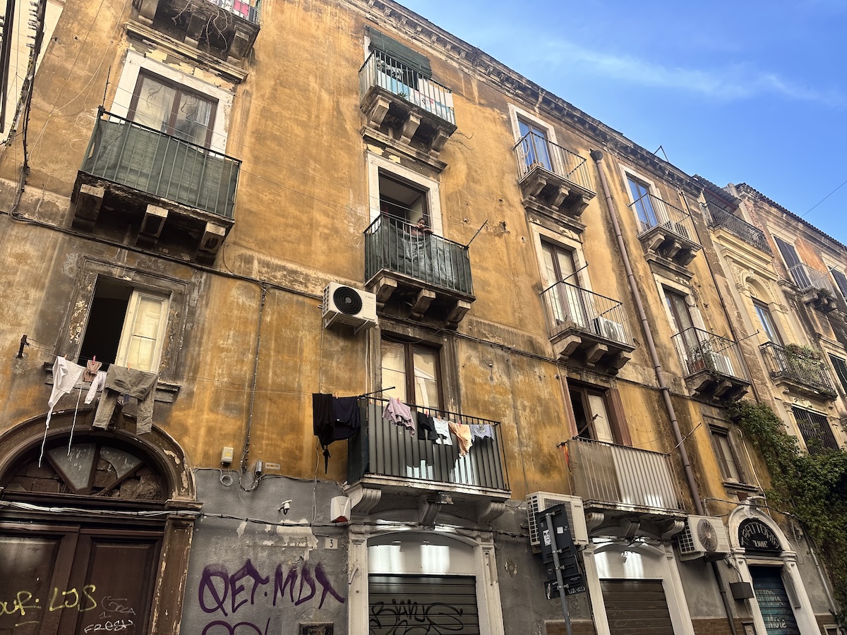 In Catania prägen raue Fassaden einer Altstadt-Gasse mit mehreren Balkonen, Wäscheleine und Graffiti das Bild, klarer Himmel im Hintergrund.