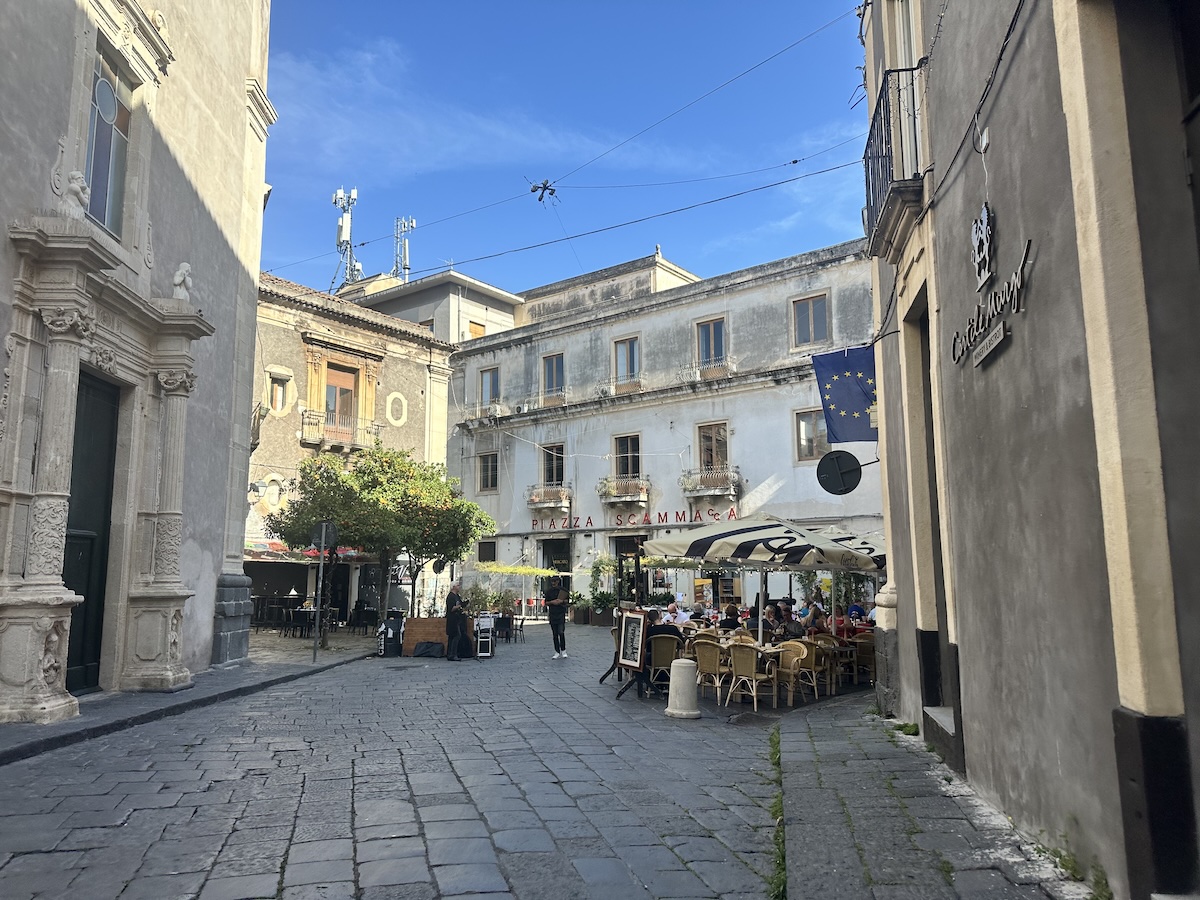 Zwischen weißen Fassaden mit blauem EU-Flaggenbanner führt Kopfsteinpflaster in eine ruhige Ecke von Catania – passend für deinen 2-Tage-Plan.