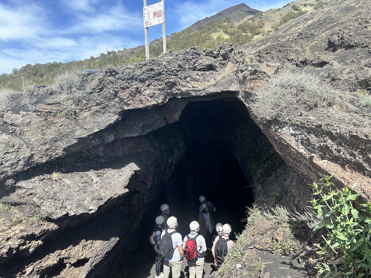 Geführte Gruppe mit Helmen vor dem dunklen Eingang einer Lavahöhle am Ätna bei Catania, umgeben von schwarzem und grauem Lavagestein.