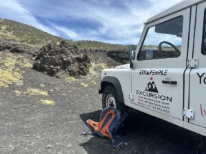 Am Ätna-Ausflug in Catania steht ein weißes EtnaPeople-Excursion Fahrzeug auf schwarzem Lavafeld, daneben ein orangefarbener Rucksack unter blauem Himmel.